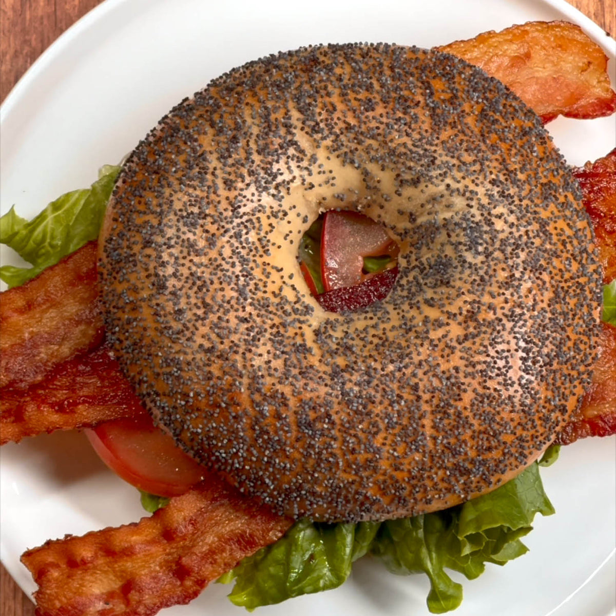 A BLT on a white plate, featuring a poppy seed bagel with crispy bacon strips, green lettuce and tomato. 